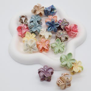 Könnte beinhalten: Eine Sammlung kleiner, blumenförmiger Haarklammern in verschiedenen irisierenden Pastellfarben wie Rosa, Blau, Grün, Gelb und Lila. Die Klammern haben ein fünfblättriges Design mit glänzender Oberfläche und sind auf einem weißen Tablett angeordnet.