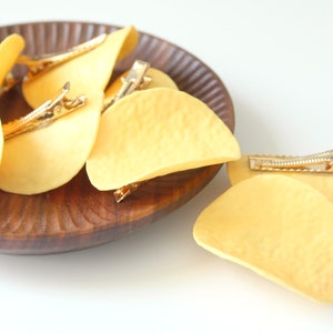 Puede incluir: Una colección de pinzas para el cabello con forma de patatas fritas amarillas. Cada pinza presenta una superficie texturizada, curva y de color amarillo claro que se asemeja a un snack, unida a un mecanismo de pinza de cocodrilo dorado.