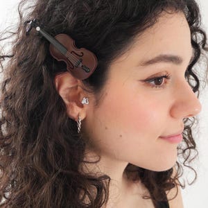 May include: A brown and black violin-shaped hair clip, a small silver-toned butterfly-shaped ear stud, and a silver-toned twisted hoop earring. The hair clip features a detailed miniature violin design with a black neck and bridge on a brown body.