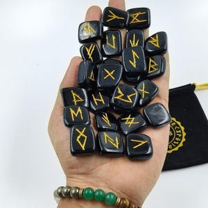 Puede incluir: Un conjunto de piedras rúnicas negras con símbolos dorados, sostenidas en una mano. Una bolsa negra con letras doradas es visible en el fondo. Una pulsera de cuentas verdes y marrones está en la muñeca.