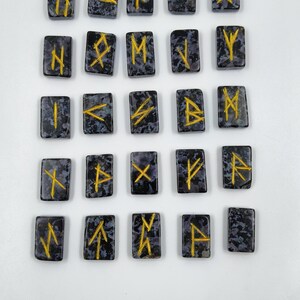 May include: A set of twenty-four rectangular rune stones. Each stone is dark gray with a speckled pattern and features a gold-colored rune symbol. The runes are arranged in a grid pattern against a white background.