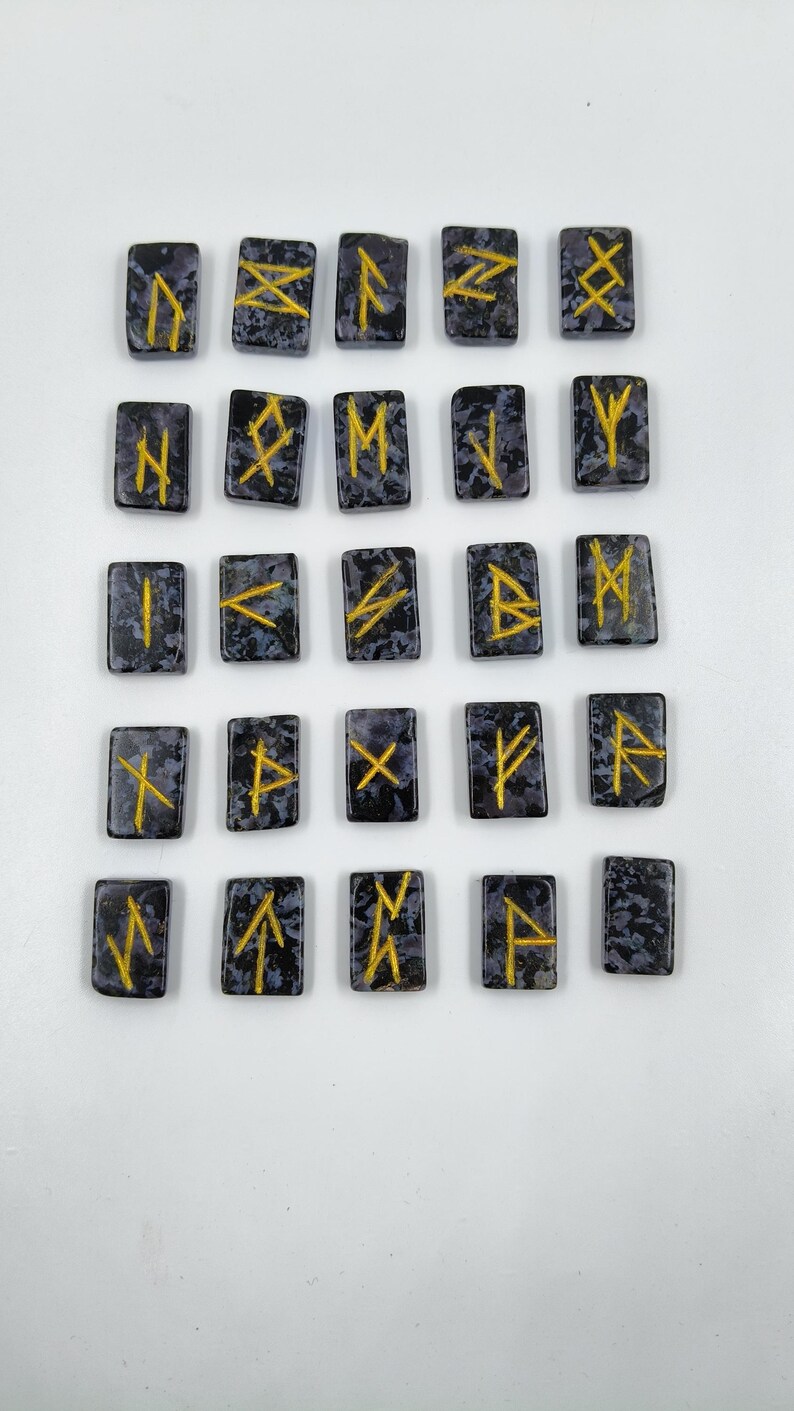 May include: A set of twenty-four rectangular rune stones. Each stone is dark gray with a speckled pattern and features a gold-colored rune symbol. The runes are arranged in a grid pattern against a white background.