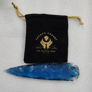 May include: A blue obsidian arrowhead with a rough, textured surface. The arrowhead is packaged in a black velvet pouch with gold embroidery that reads "Tashada Export The Crystal Shop".