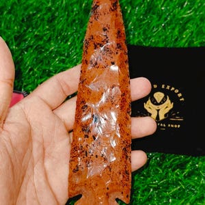 May include: A large, reddish-brown arrowhead with a pointed tip and a slightly curved base. The arrowhead has a translucent, almost glassy appearance, with flecks of black throughout.