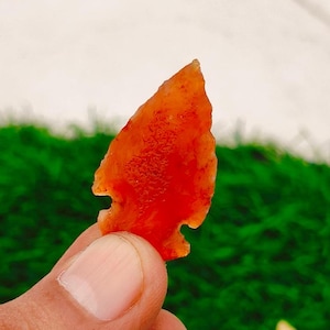May include: A translucent, orange arrowhead with a textured surface. The arrowhead is held between two fingers against a backdrop of green and white.