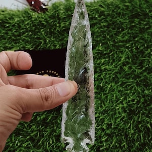 Puede incluir: Una punta de flecha transparente hecha de vidrio. La punta de flecha es puntiaguda en la parte superior y tiene una base redondeada. Mide aproximadamente 15 cm de largo.