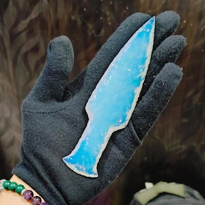 Puede incluir: Una punta de flecha de vidrio de color azul claro iridiscente. El vidrio es translúcido y tiene una superficie lisa y pulida.