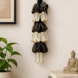 May include: A hanging wall decor piece featuring alternating black and white stones encased in gold-colored wire. The stones are suspended from a black rope with a green accent. The decor is displayed against a neutral wall, with a plant and books in the foreground.