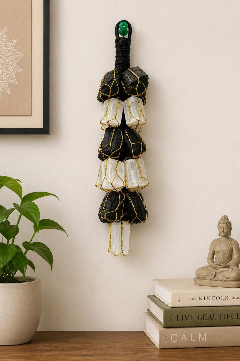 May include: A hanging wall decor piece featuring alternating black and white stones encased in gold-colored wire. The stones are suspended from a black rope with a green accent. The decor is displayed against a neutral wall, with a plant and books in the foreground.