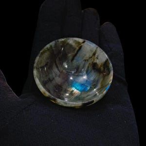 May include: A small, round bowl made of labradorite, a gemstone known for its iridescent blue and green hues. The bowl is smooth and polished, with a natural, organic shape.