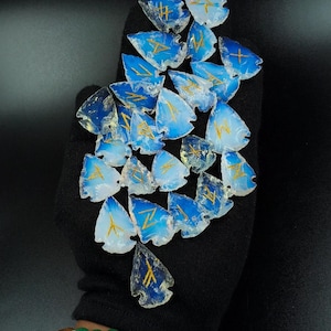 May include: A collection of blue and white arrowhead-shaped pendants with gold-coloured symbols. The pendants are held in a black gloved hand. A beaded bracelet with green and gold beads is visible.
