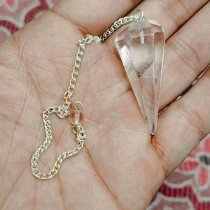 Könnte beinhalten: Ein klares Quarzkristall-Pendel mit einer silbernen Kette und einer kleinen durchsichtigen Perle. Das Pendel wird in einer Hand gehalten.