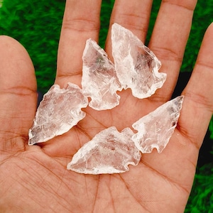 Puede incluir: Cinco puntas de flecha de cuarzo transparente en una mano. Los cristales son translúcidos y tienen una textura natural y rugosa.