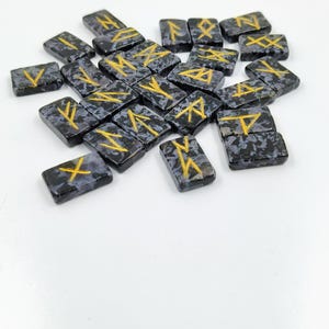 May include: A collection of rectangular rune stones. Each stone is dark gray with a speckled pattern and features a gold-colored rune symbol. The stones are scattered on a white surface.