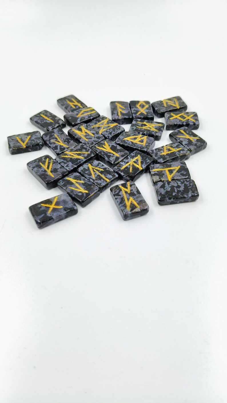 May include: A collection of rectangular rune stones. Each stone is dark gray with a speckled pattern and features a gold-colored rune symbol. The stones are scattered on a white surface.