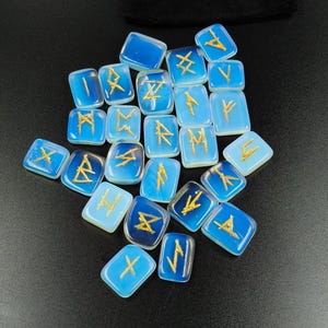 Peut inclure: Ensemble de pierres runiques bleues avec des symboles dorés, disposées sur une surface noire. Un sac en velours noir avec l'inscription dorée "RUNE STONES" est visible. Les pierres rectangulaires varient en nuances de bleu.