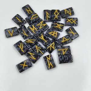May include: A collection of rectangular rune stones with a speckled, dark gray background and gold-colored symbols. The runes are scattered across a white surface, offering a glimpse into ancient divination practices.