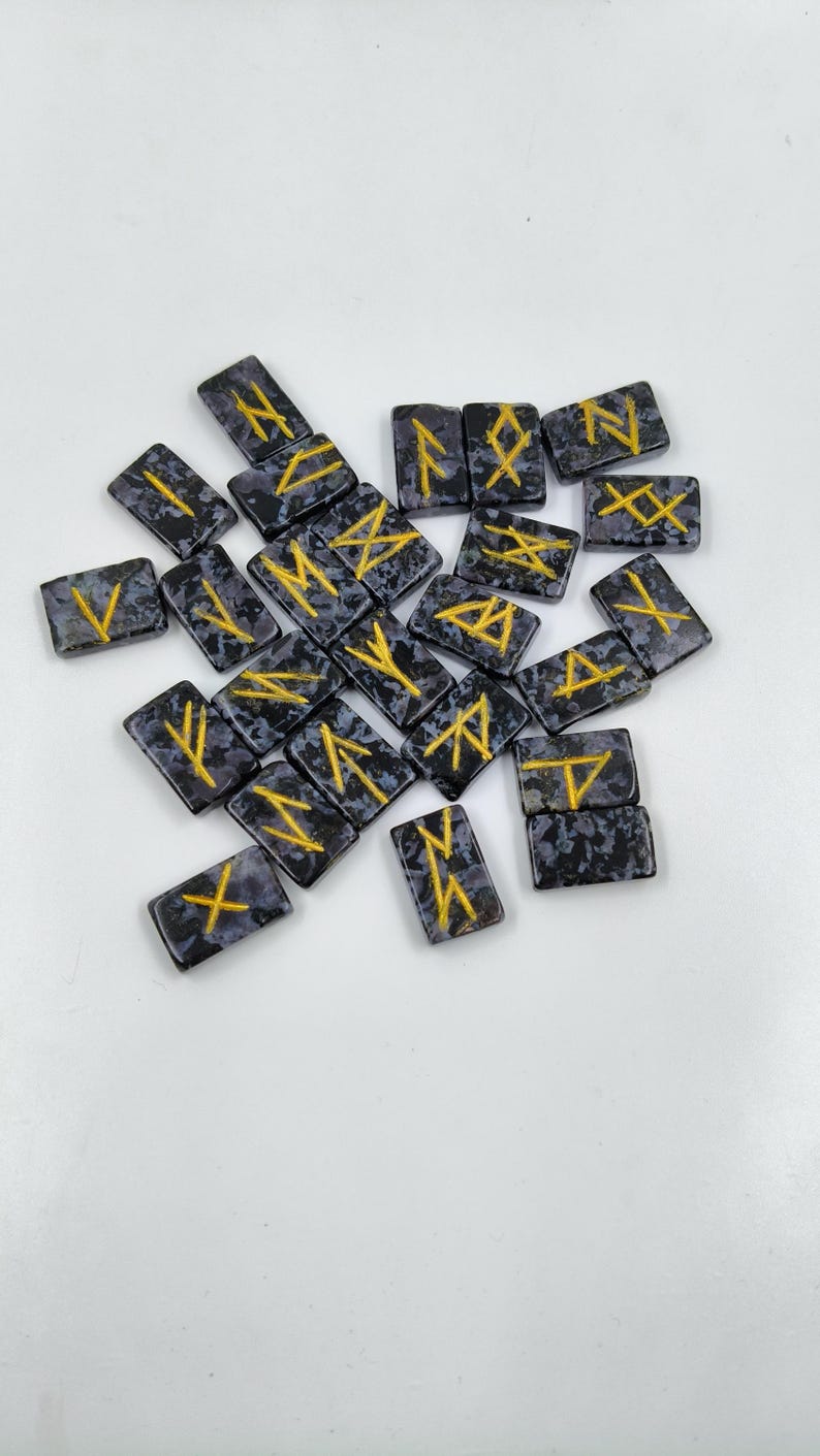 May include: A collection of rectangular rune stones with a speckled, dark gray background and gold-colored symbols. The runes are scattered across a white surface, offering a glimpse into ancient divination practices.