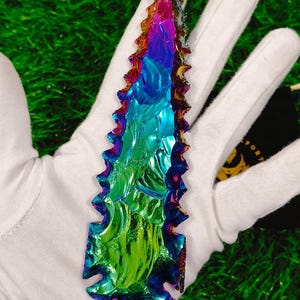 May include: A rainbow iridescent obsidian arrowhead with a serrated edge. The arrowhead is held in a white glove against a green background.