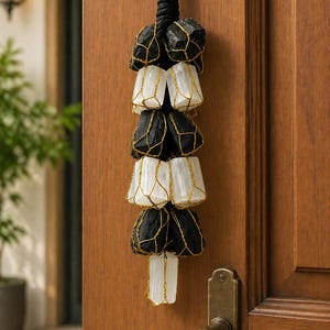 May include: A decorative hanging ornament featuring alternating black and white stones encased in gold-colored wire. The ornament is suspended from a black rope and hangs against a wooden door.