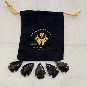 Puede incluir: Cinco puntas de flecha de obsidiana negra en una bolsa de terciopelo con cordón de ajuste dorado. La bolsa tiene el texto "Tasha Export The Crystal Shop" impreso en ella.