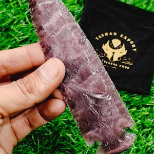 May include: A purple and gray arrowhead made of natural stone. The arrowhead is held in a hand and is resting on a bed of green grass. The arrowhead is about 6 inches long.