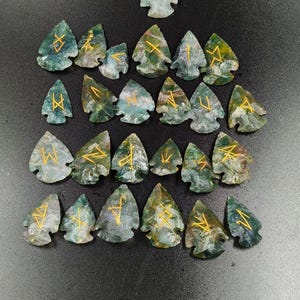 Może przedstawiać: Kolekcja kamieni runicznych z mchu agatowego, każdy w kształcie grotu strzały. Kamienie są mieszanką zieleni i bieli ze złotymi symbolami runicznymi. Tło to ciemna, teksturowana powierzchnia.