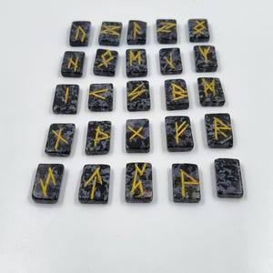 May include: A set of twenty-four rectangular rune stones. Each stone is dark gray with a speckled pattern and features a gold-colored rune symbol. The stones are arranged on a white surface.