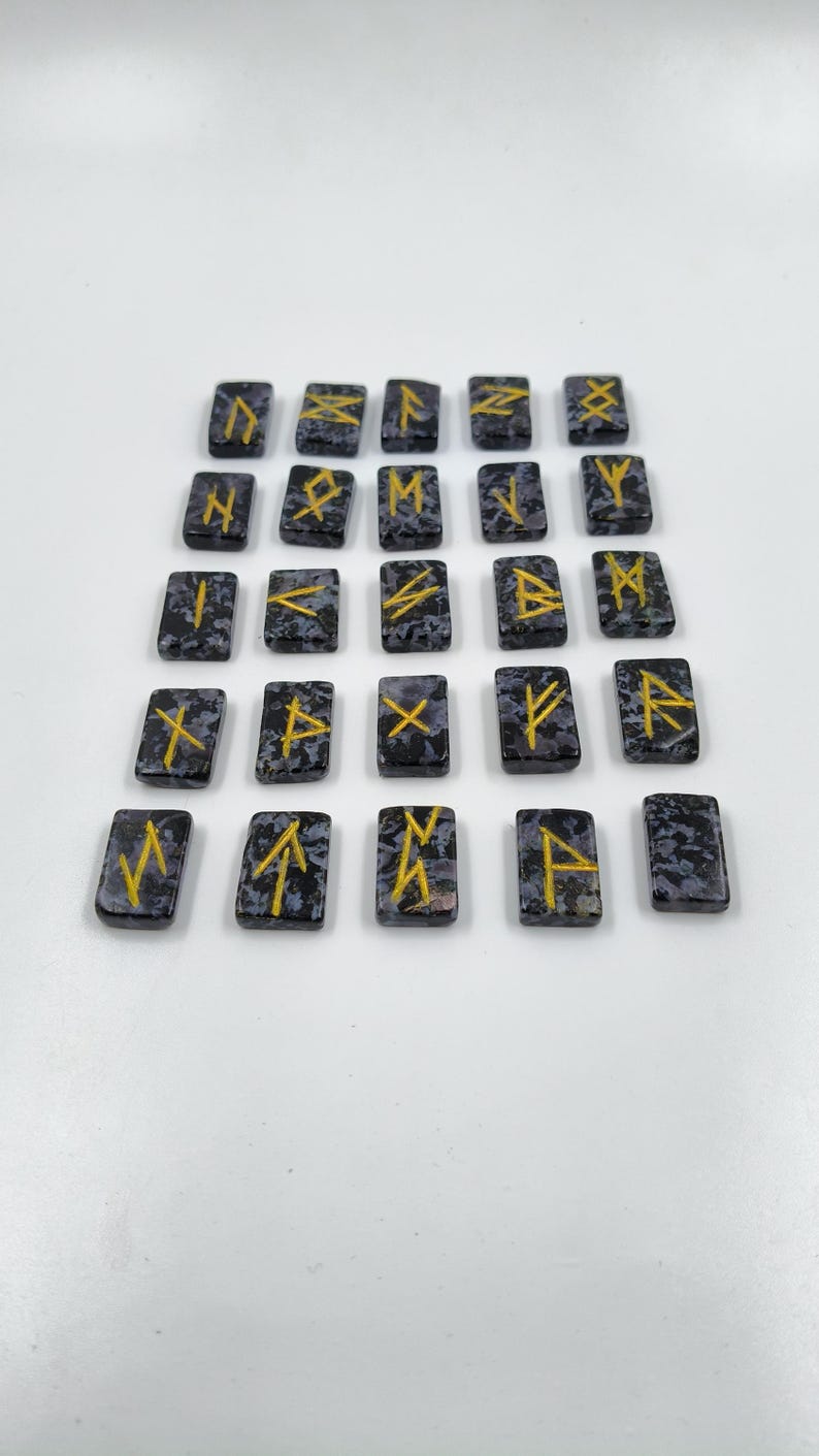 May include: A set of twenty-four rectangular rune stones. Each stone is dark gray with a speckled pattern and features a gold-colored rune symbol. The stones are arranged on a white surface.