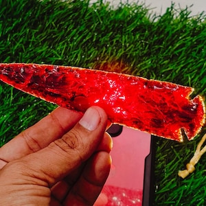 May include: A hand holding a translucent, red arrowhead. The arrowhead has a sharp point and a notched base. The background is green, with a small, tan, twisted cord visible.