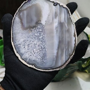 May include: A white and grey agate slice with a silver trim. The slice is approximately 12.5 cm in diameter and has a natural, organic shape.