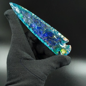 May include: A hand in a black glove holds a blue and green glass arrowhead. The arrowhead has a sharp point and a textured surface. The colours shift in the light, creating an iridescent effect.