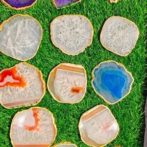 May include: A collection of agate coasters with gold trim. The coasters are various colours, including purple, white, orange, blue, and green. The coasters are arranged on a green grassy surface.