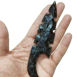 May include: A black obsidian arrowhead with a serrated edge and a curved base. The arrowhead is held in a hand against a white background. The obsidian has a glossy, reflective surface.