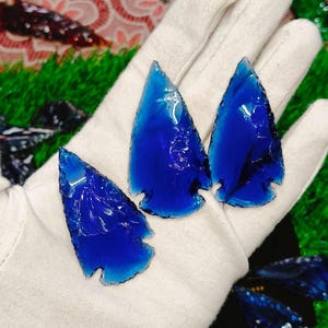 May include: Four vibrant blue glass arrowhead replicas are displayed on a white glove. The arrowheads have a translucent quality, with sharp edges and a classic arrowhead shape. The background includes green and black elements.