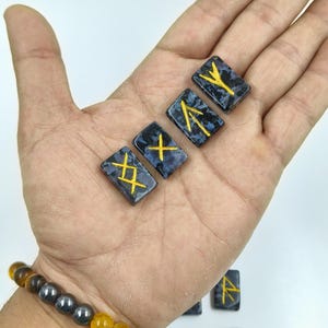 May include: A hand displays several rectangular, dark blue rune stones with gold-colored symbols. The stones have a speckled pattern. A beaded bracelet with yellow, purple, and gray beads is visible on the wrist.