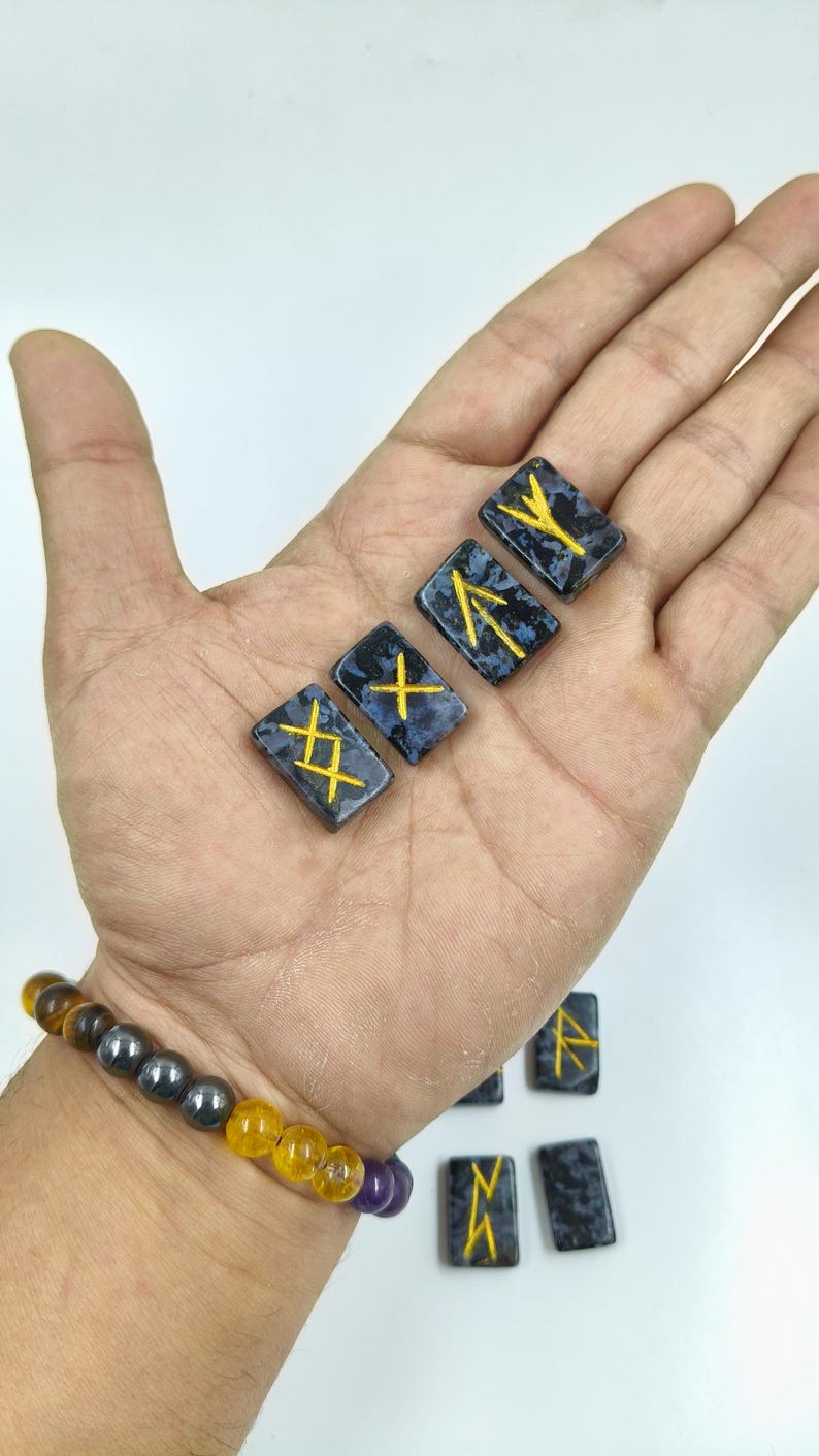 May include: A hand displays several rectangular, dark blue rune stones with gold-colored symbols. The stones have a speckled pattern. A beaded bracelet with yellow, purple, and gray beads is visible on the wrist.