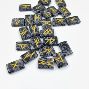 May include: A collection of rectangular, dark gray rune stones with gold-colored symbols. The stones have a speckled pattern and are scattered on a white surface. The symbols are various geometric shapes and lines.