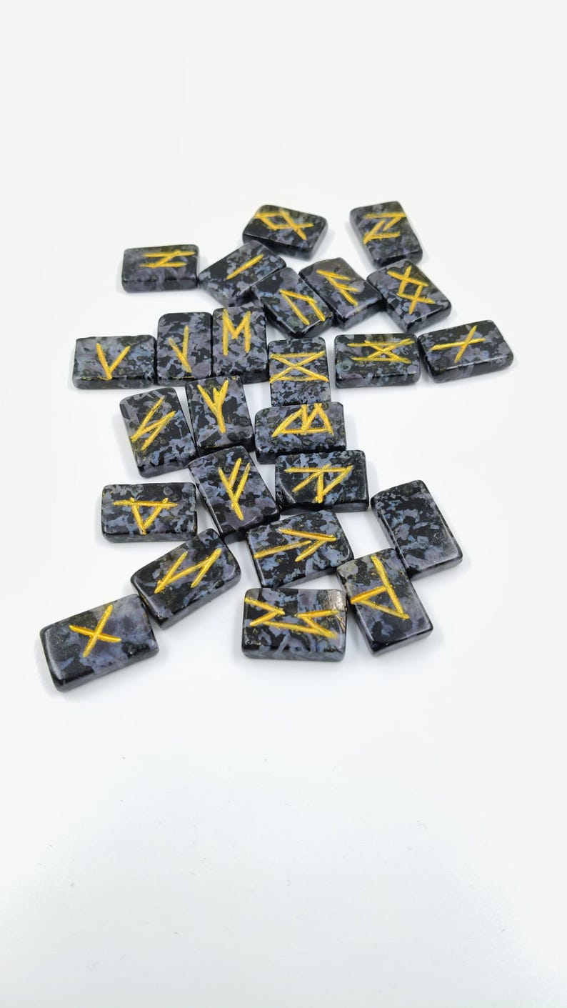 May include: A collection of rectangular, dark gray rune stones with gold-colored symbols. The stones have a speckled pattern and are scattered on a white surface. The symbols are various geometric shapes and lines.