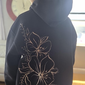 Puede incluir: Una sudadera con capucha negra que presenta un gran diseño de arte lineal floral de color claro en la espalda. El gráfico consta de tres flores prominentes con tallos delicados, hojas y pequeños acentos punteados, en un tono beige suave y apagado.