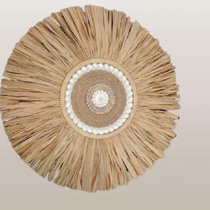 Op de afbeelding: Cirkelvormige wanddecoratie gemaakt van natuurlijke geweven materialen. Het ontwerp heeft een centrale geweven cirkel, een ring van witte kralen en een franje van gedroogde bladeren. Het kleurenpalet is neutraal, met tinten beige en bruin.