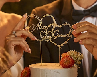 May include: A two-tiered wedding cake with white frosting and a gold heart-shaped cake topper that says "Alice & Samuel 28.24".