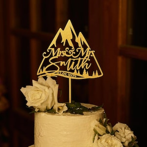 Peut inclure: Un gâteau de mariage à deux étages décoré de glaçage blanc et de verdure. Un topper de gâteau en forme de montagne dorée avec le texte "Mr. & Mrs. Smith 02.05.2023" est placé sur le dessus du gâteau.