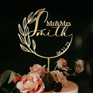 May include: Gold wedding cake topper with a leafy wreath and the text "Mr & Mrs Smith 06.21.23".
