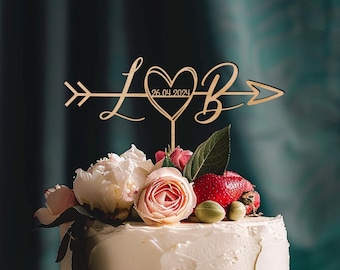 May include: Wooden wedding cake topper with a heart and arrow design. The heart has the letters "L" and "B" on either side and the date "26.04.2024" inside. The topper is decorated with pink and white flowers.