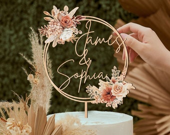 Puede incluir: Un adorno para pastel de madera con un marco circular y los nombres "James & Sophia" escritos en cursiva. El marco está decorado con un diseño floral en tonos marrones, naranjas y blancos.