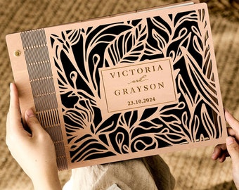 May include: A wooden photo album with a floral design cut out. The album is light brown with a black floral pattern. The text "VICTORIA and GRAYSON 23.10.2024" is printed in gold on the front.