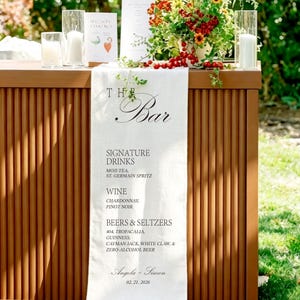 May include: A white fabric bar menu runner draped over a brown fluted bar. The text reads THE Bar, SIGNATURE DRINKS, MOJI-TEA, ST. GERMAIN SPRITZ, WINE, CHARDONNAY, PINOT NOIR, BEERS & SELTZERS, 404, TROPACALIA, GUINNESS, CAYMAN JACK, WHITE CLAW, & ZERO-ALCOHOL BEER, Angela + Simon, 02. 21. 2026.