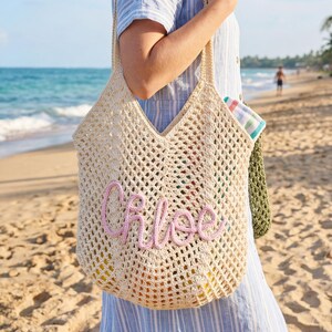 May include: A beige crocheted tote bag with the name "Chloe" embroidered in pink script. The bag has a large, open weave design and is carried over the shoulder. The background shows a beach scene with the ocean and palm trees.