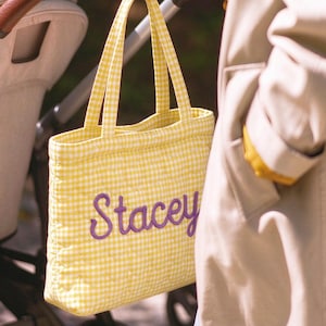 May include: A yellow and white gingham quilted tote bag featuring the name Stacey embroidered in purple script lettering. The bag is suspended from a silver-toned metal hook attached to the leather-wrapped handle of a stroller.